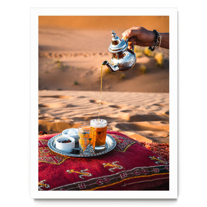 A hand pours tea from a silver teapot into two glasses on a tray, set on a red and gold patterned cushion, with a desert landscape in the background.