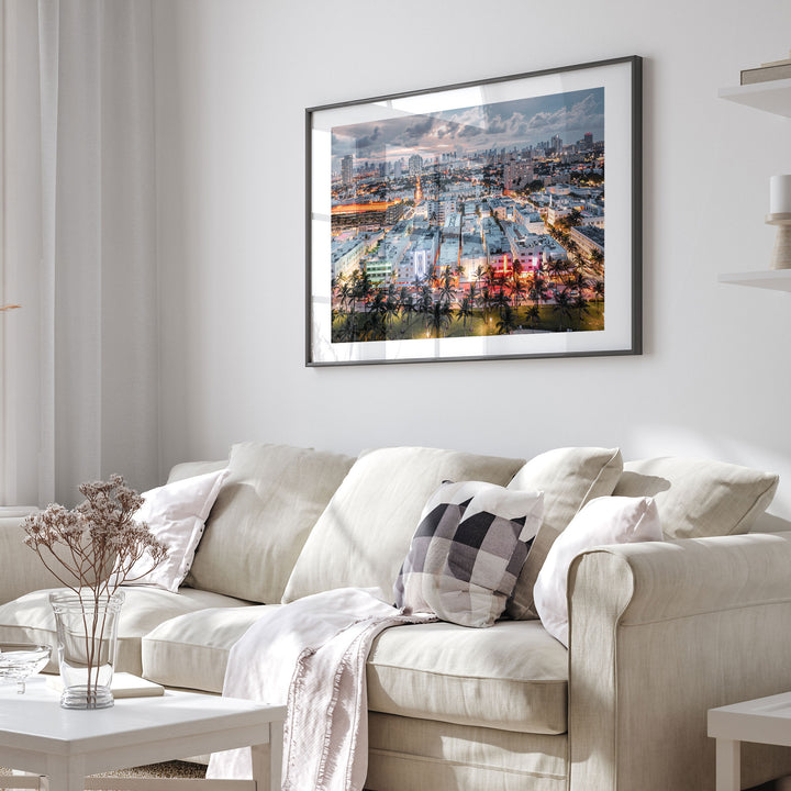 a cozy living room with a beige sofa adorned with a checkered throw pillow and a white blanket draped over it. Above the sofa, a large framed picture of a cityscape with a mix of buildings and trees is displayed on the wall.