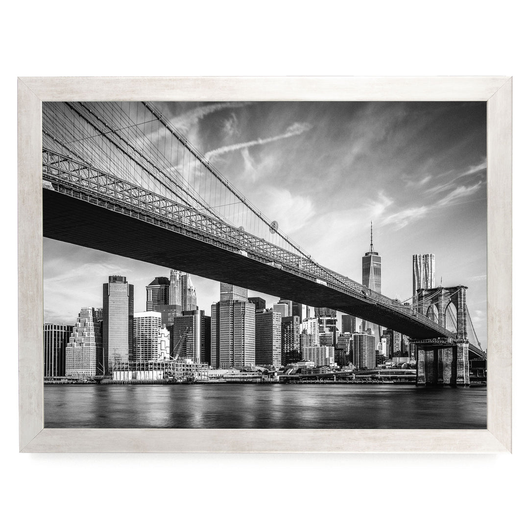 A black and white photograph of the Brooklyn Bridge spanning the East River, with the New York City skyline visible in the background.