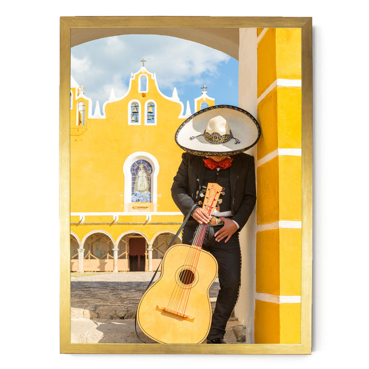 A person dressed in a traditional Mexican outfit, including a sombrero, is holding a guitar and leaning against a yellow building with arched windows.