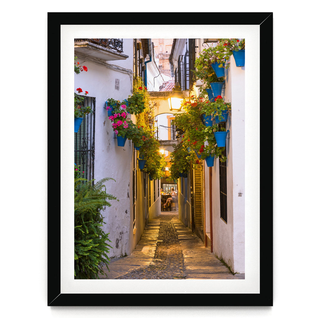 A narrow cobblestone street lined with colorful flower pots and plants, leading to a building with a balcony and a person sitting on a bench.