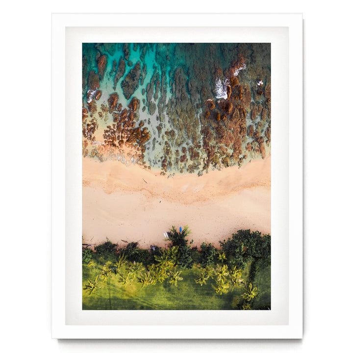 The image is a framed aerial view of a beach with a mix of sandy and rocky areas, surrounded by lush greenery and palm trees.