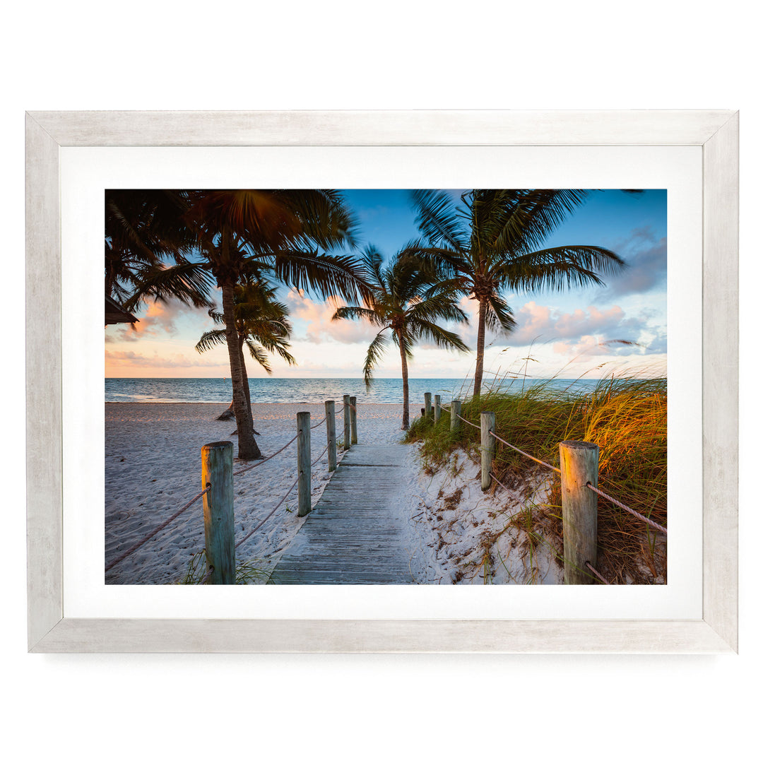 A wooden walkway leads to a beach lined with palm trees, with the ocean visible in the distance.