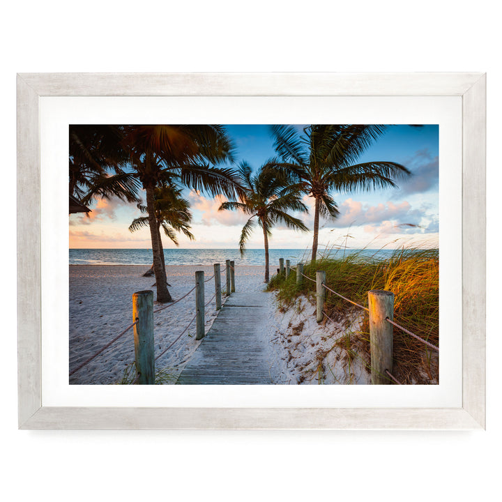 A wooden walkway leads to a beach lined with palm trees, with the ocean visible in the distance.