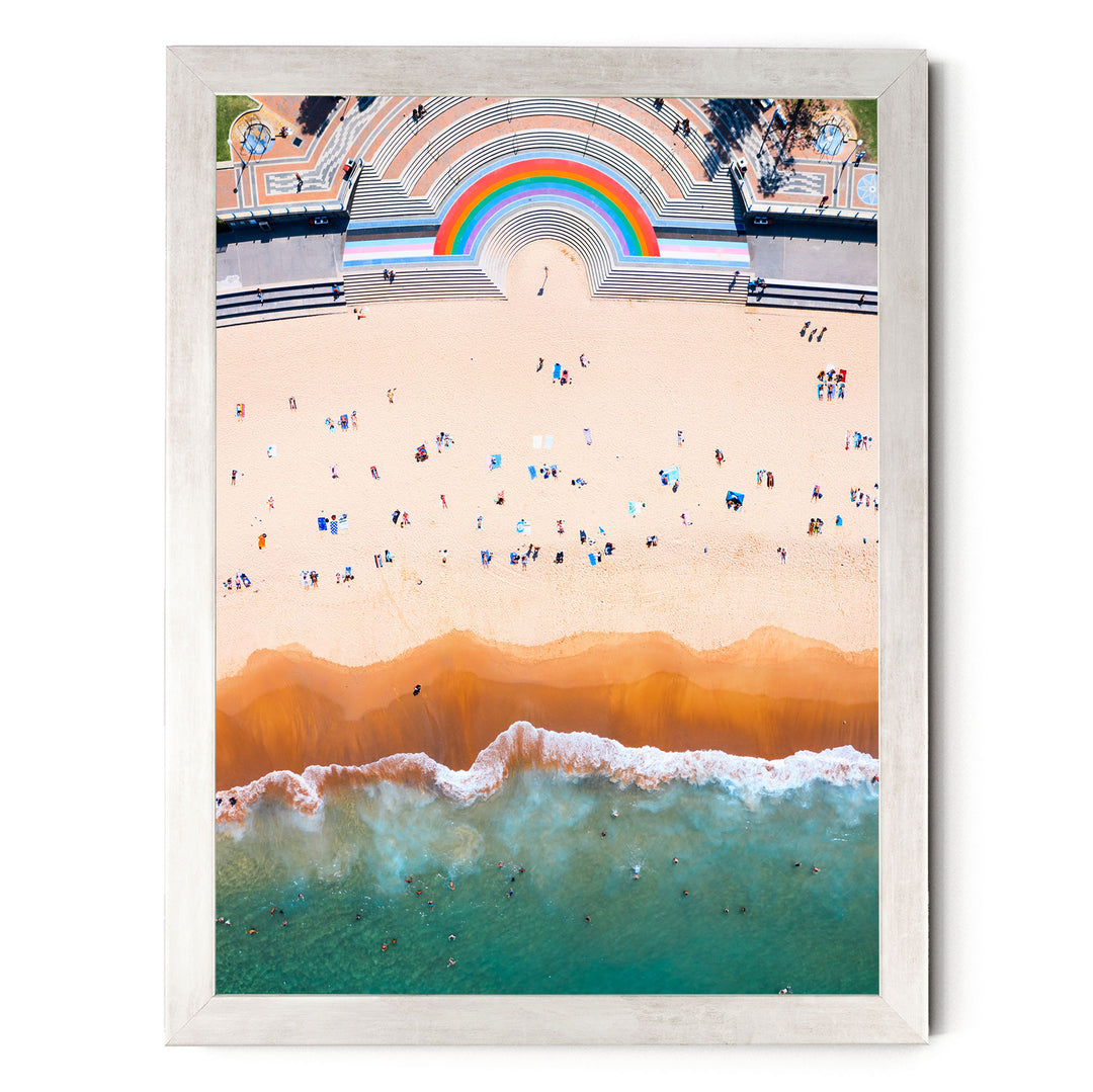 The image is an aerial view of a beach with a large rainbow arching over the top, and people enjoying the sandy shoreline.
