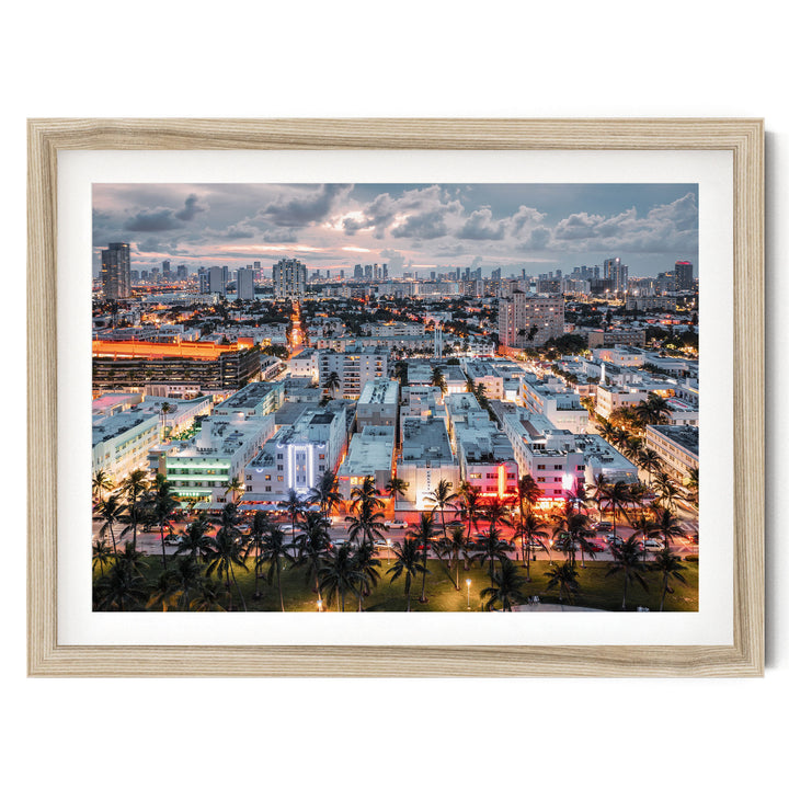 The image is a vibrant cityscape at dusk, with a skyline of tall buildings and skyscrapers illuminated by colorful lights. Palm trees line the streets, adding a tropical touch to the urban landscape. The scene is captured from an aerial perspective, providing a comprehensive view of the city's architectural diversity and the interplay of light and shadow.