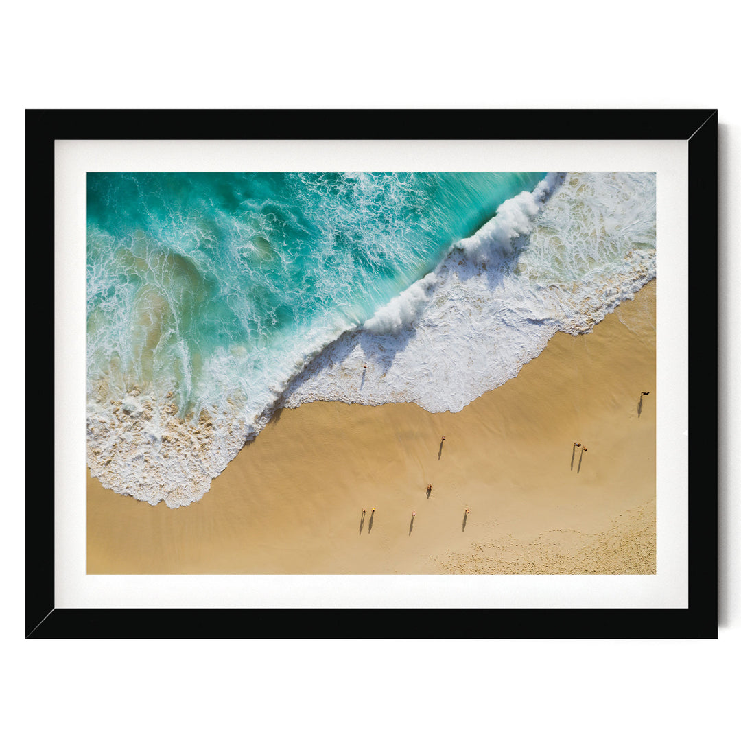 The image is a framed aerial view of a beach with people walking along the shoreline, where the waves are crashing onto the sand.