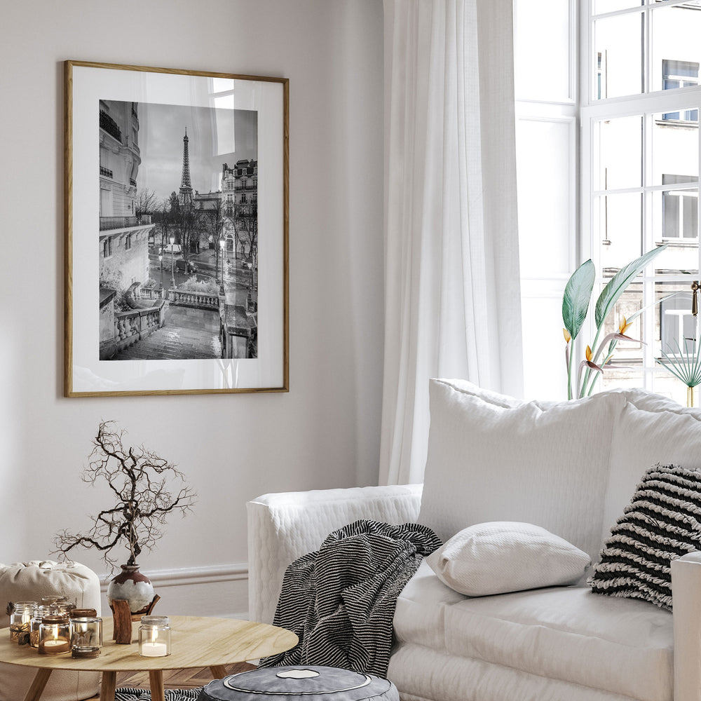 a cozy living room with a large framed black and white photograph of the Eiffel Tower hanging on the wall. The room is decorated with a white sofa, a wooden coffee table, and various decorative items such as a vase, a plant, and a striped throw blanket.