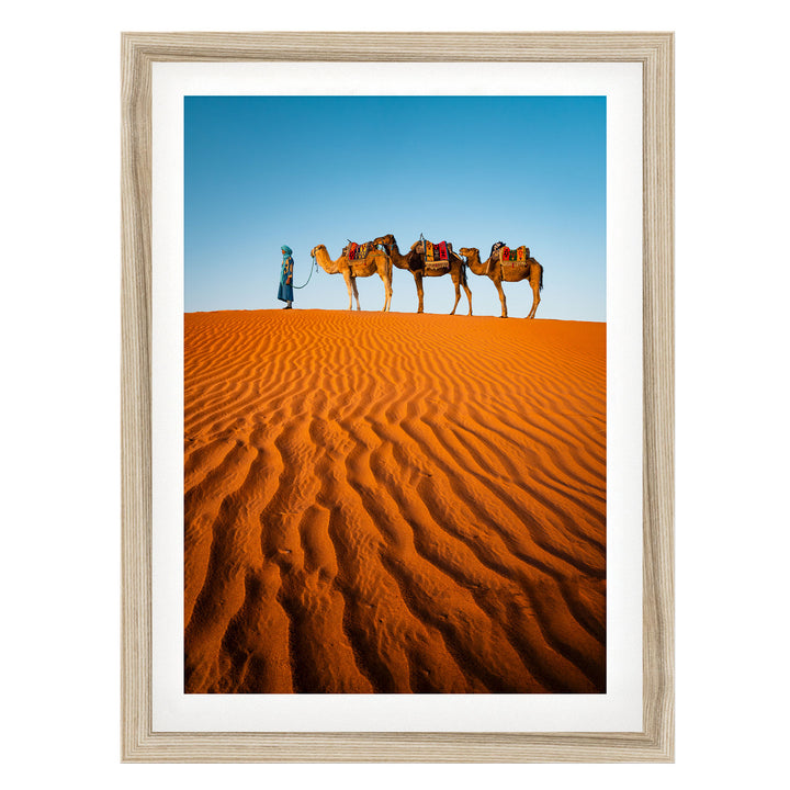 A person in a blue robe leads three camels across a vast, orange sand dune in the desert.