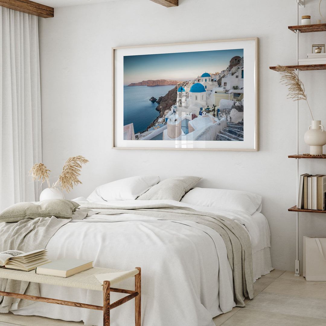 a bedroom with a large framed picture of a scenic view of a coastal town with white buildings and blue roofs, hanging on the wall above a bed with white linens and a gray blanket.