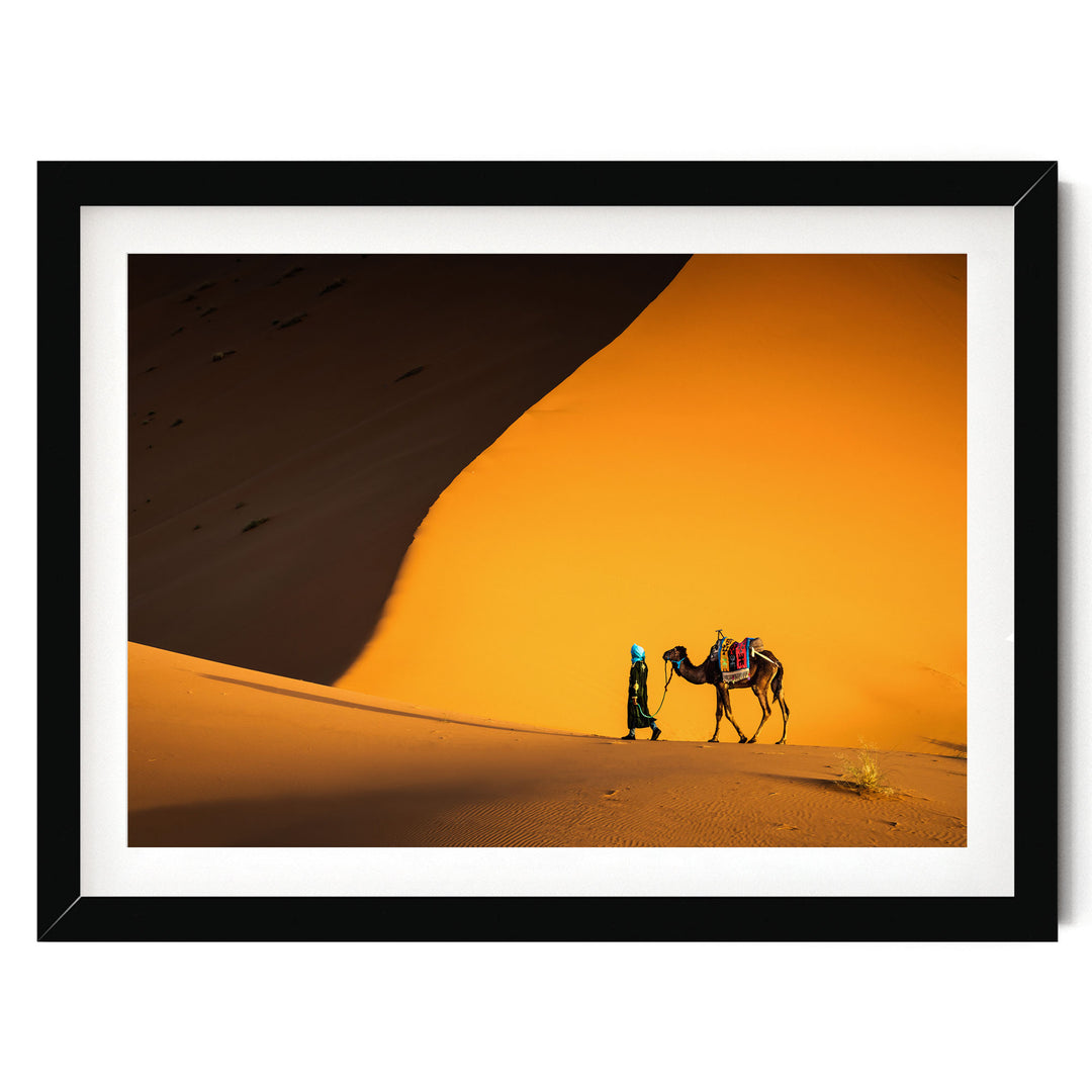 A person is walking a camel across a desert landscape with a large sand dune in the background.
