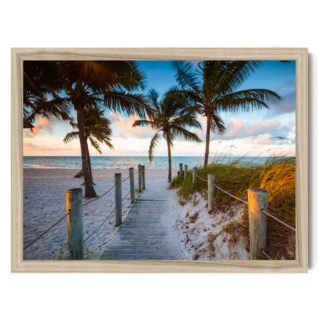 A wooden walkway leads to a tropical beach, with swaying palm trees and a clear blue sky in the background.