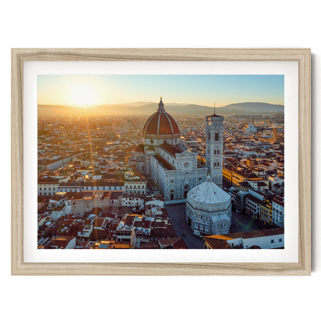 a cityscape with a large domed building, possibly a cathedral or church, surrounded by smaller buildings and structures. The sun is setting, casting a warm glow over the scene and creating a picturesque view of the city.