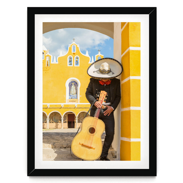 A person dressed in traditional Mexican attire, including a sombrero and mariachi outfit, is playing a guitar in front of a yellow building with arched windows.