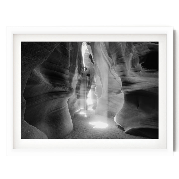 A black and white photograph of a narrow passage in a rock formation, with sunlight streaming through the opening and casting shadows on the walls.