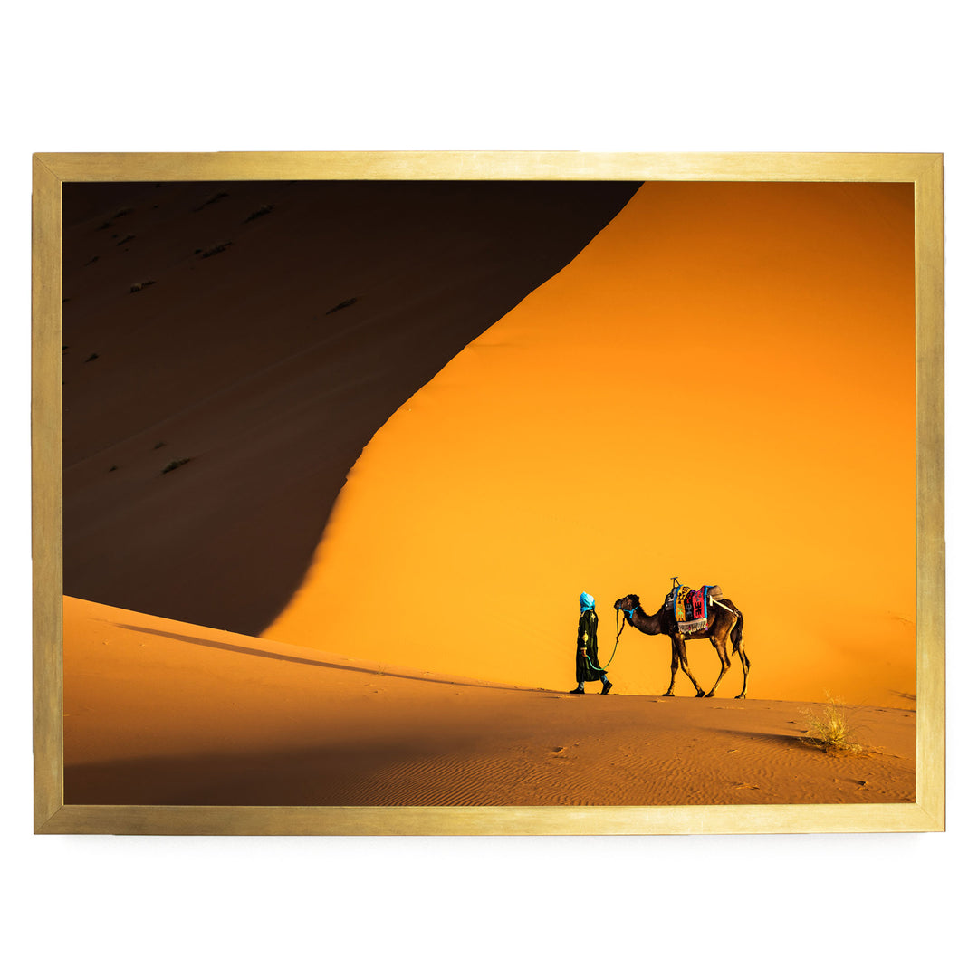 A person in a green outfit is walking a camel across a sandy desert landscape, with a large sand dune in the background.