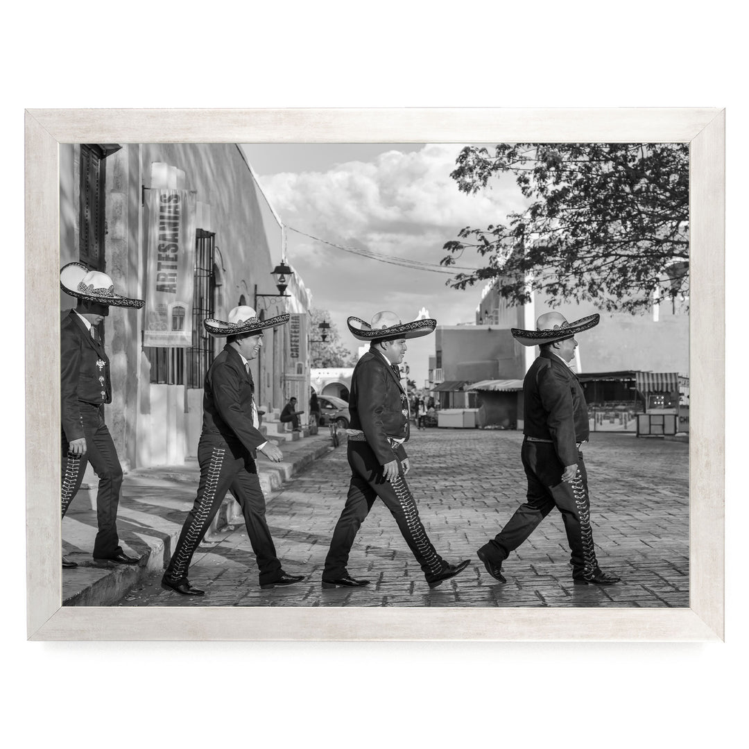 A group of men wearing sombreros and dark clothing are walking down a street, with a building and trees visible in the background.
