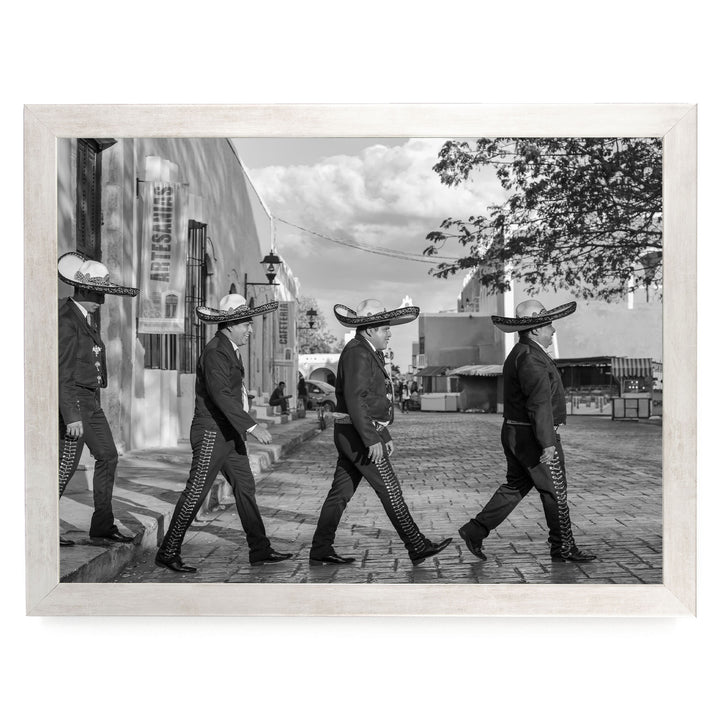 A group of men wearing sombreros and dark clothing are walking down a street, with a building and trees visible in the background.