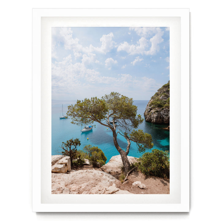 A scenic view of a rocky coastline with a large tree, a small sailboat, and a larger boat in the distance, all set against a backdrop of a blue sky with clouds.