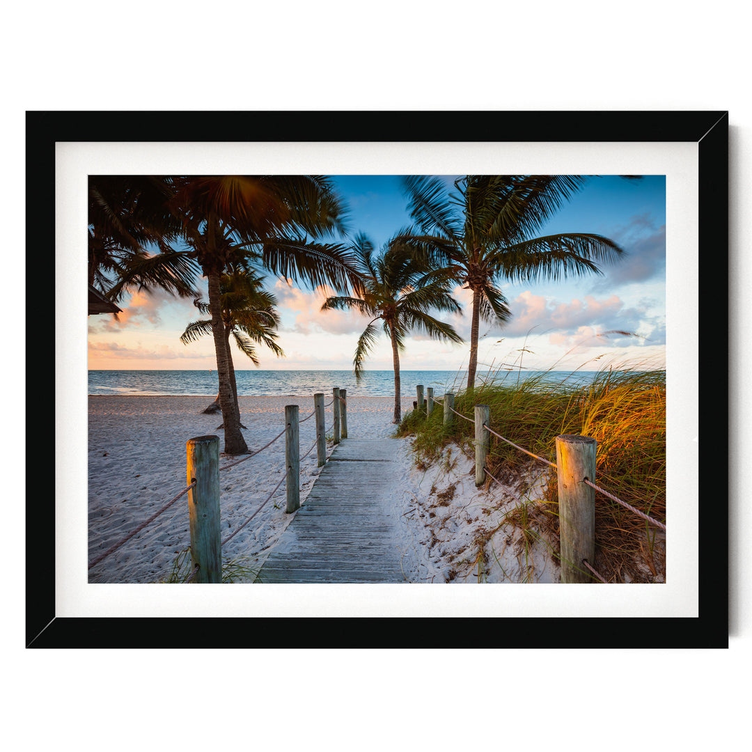 A wooden walkway leads to a sandy beach, with palm trees lining the path and a colorful sunset sky in the background.