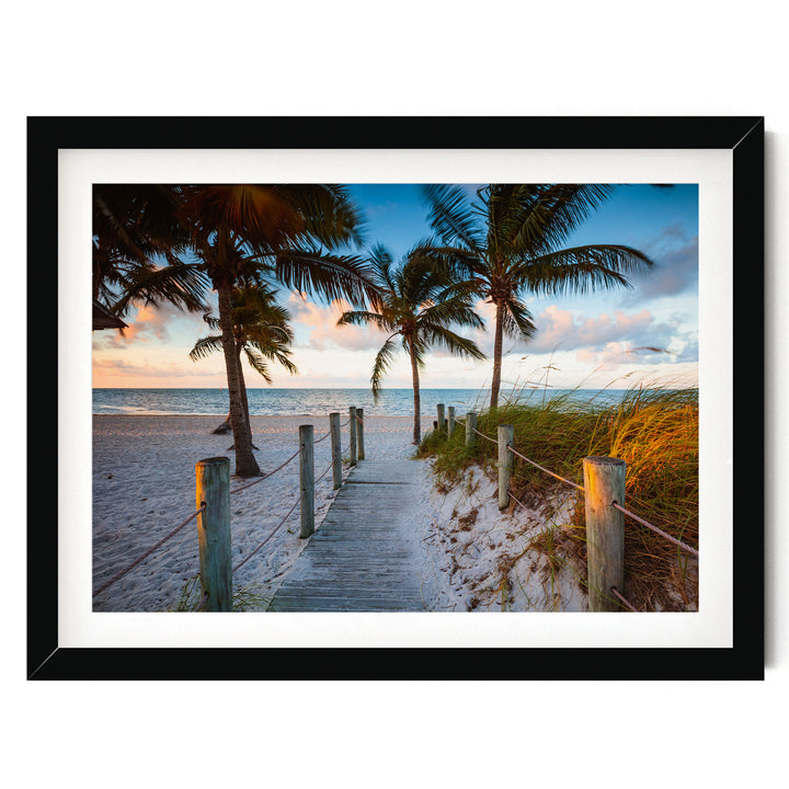 A wooden walkway leads to a sandy beach, with palm trees lining the path and a colorful sunset sky in the background.