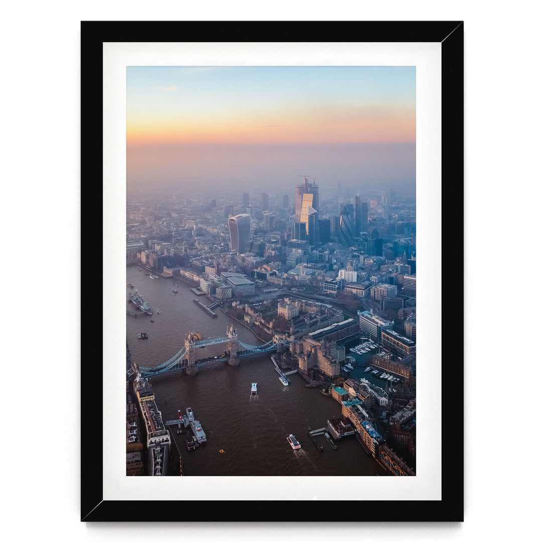 The image is an aerial view of a cityscape with a river running through it, featuring a bridge and several boats. The city is densely populated with tall buildings, and the sky is filled with warm, hazy colors, suggesting a sunrise or sunset.