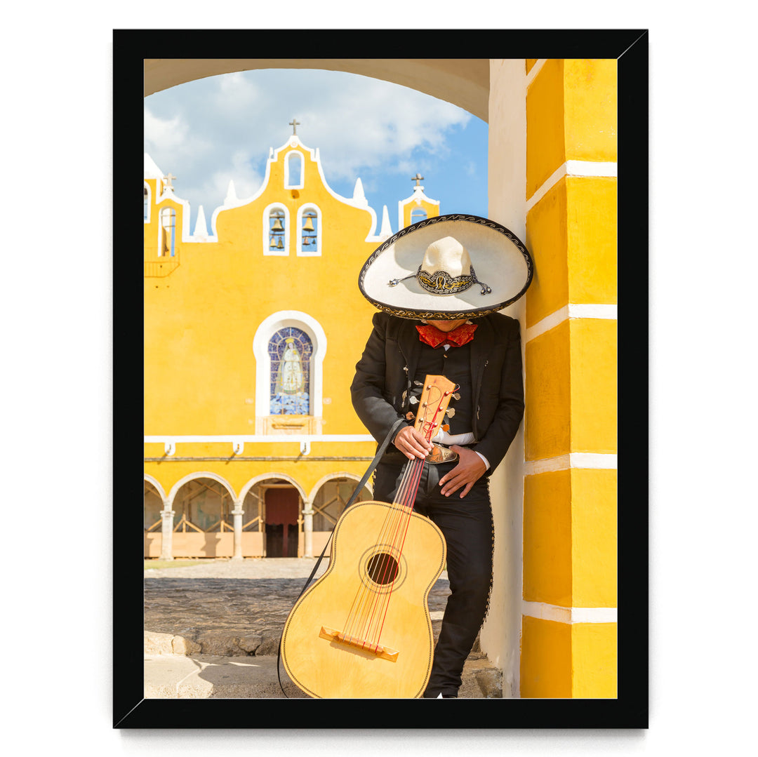 A person in a sombrero and traditional Mexican attire is playing a guitar in front of a yellow building with arched windows and a cross on top.
