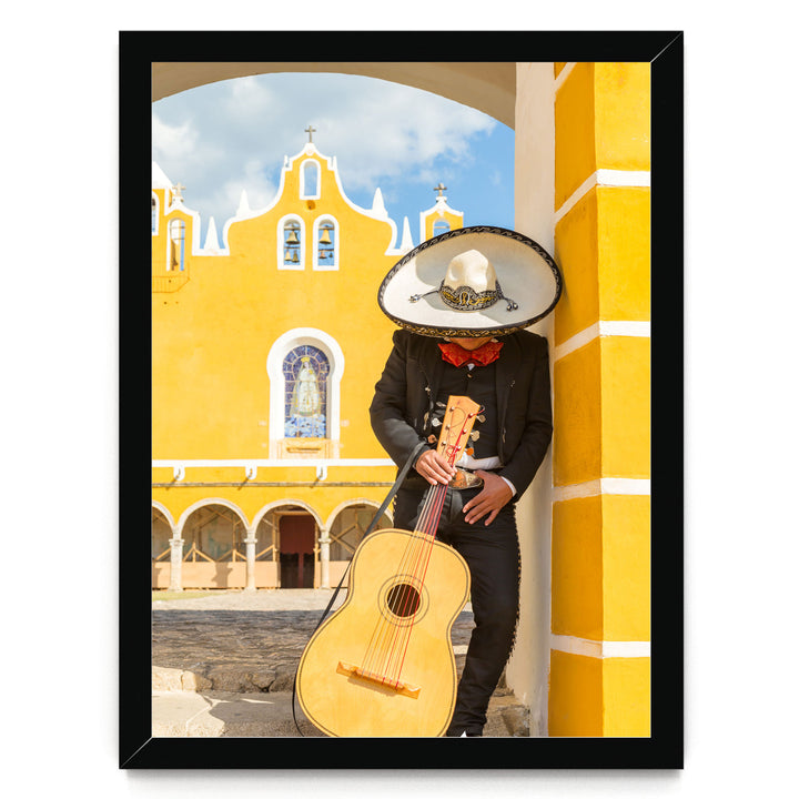 A person in a sombrero and traditional Mexican attire is playing a guitar in front of a yellow building with arched windows and a cross on top.