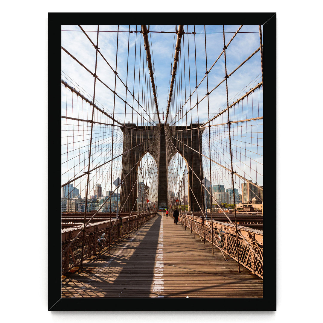 the iconic Brooklyn Bridge, with its distinctive Gothic-style towers and suspension cables, stretching across the frame. The bridge is framed within a black border, and a person can be seen walking on the wooden deck. The scene is set against a backdrop of a city skyline, creating a striking contrast between the architectural marvel and the urban landscape.