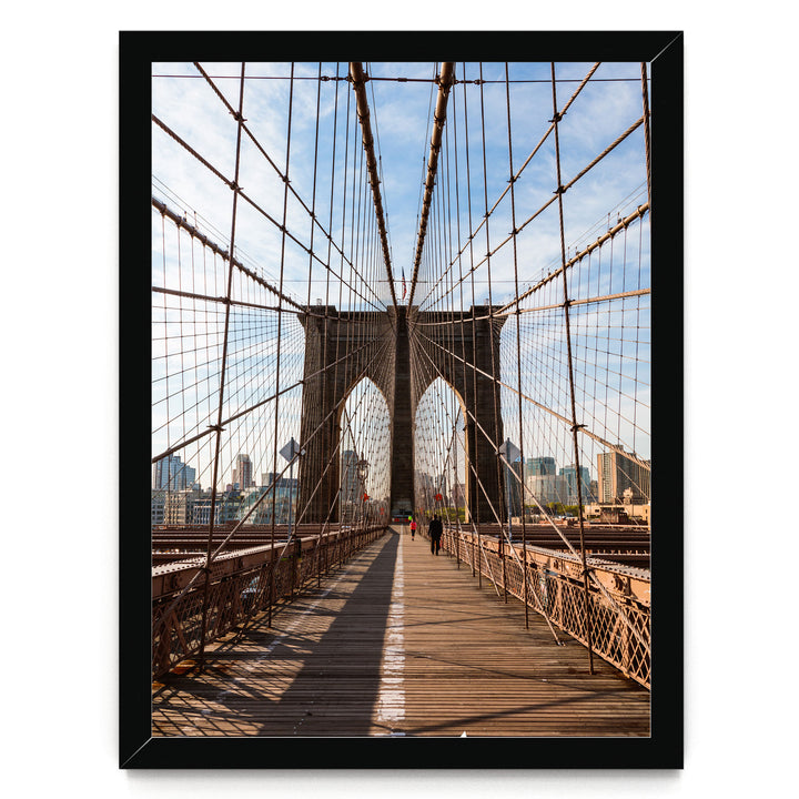 the iconic Brooklyn Bridge, with its distinctive Gothic-style towers and suspension cables, stretching across the frame. The bridge is framed within a black border, and a person can be seen walking on the wooden deck. The scene is set against a backdrop of a city skyline, creating a striking contrast between the architectural marvel and the urban landscape.