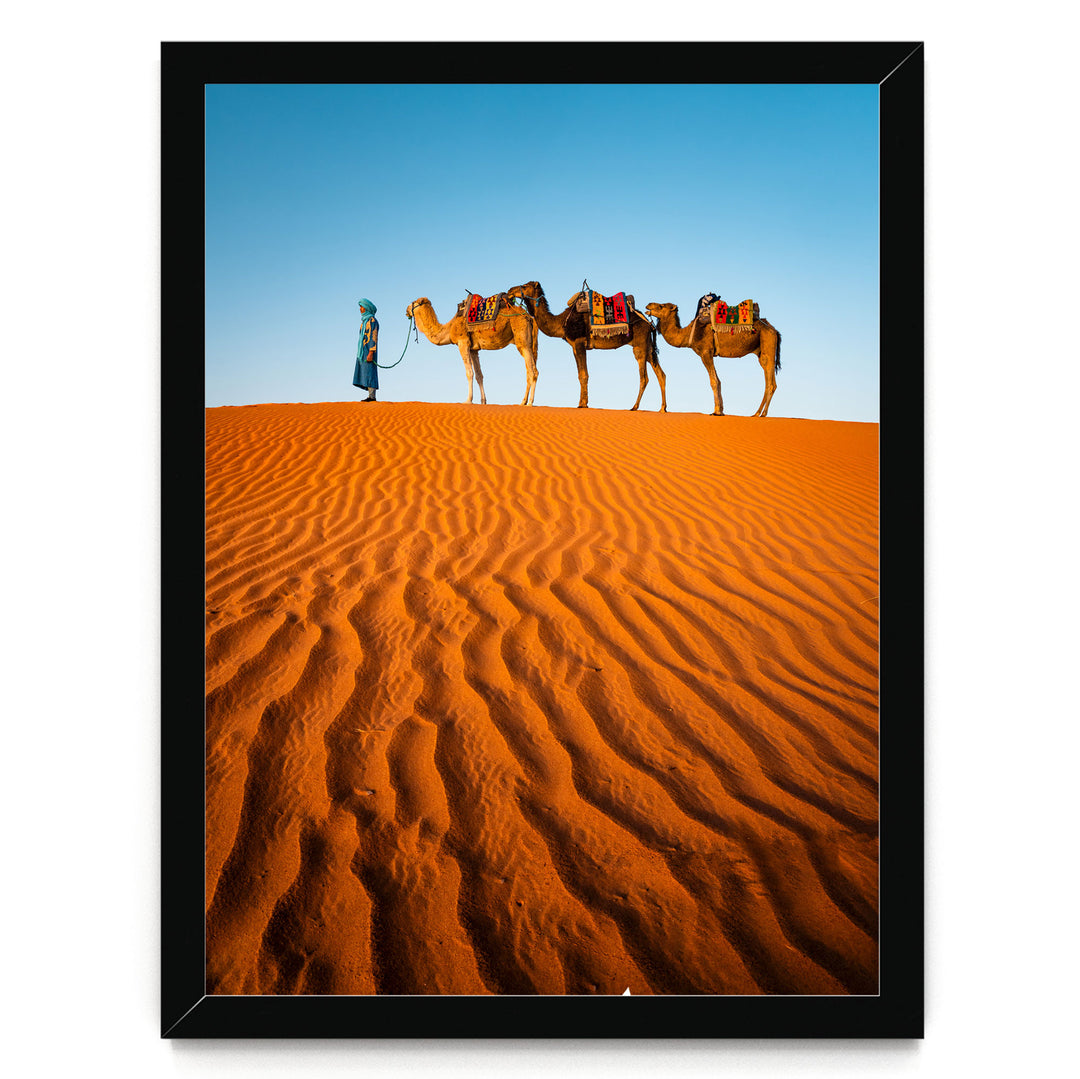 A person in a blue robe leads three camels across a vast, undulating sand dune under a clear blue sky.