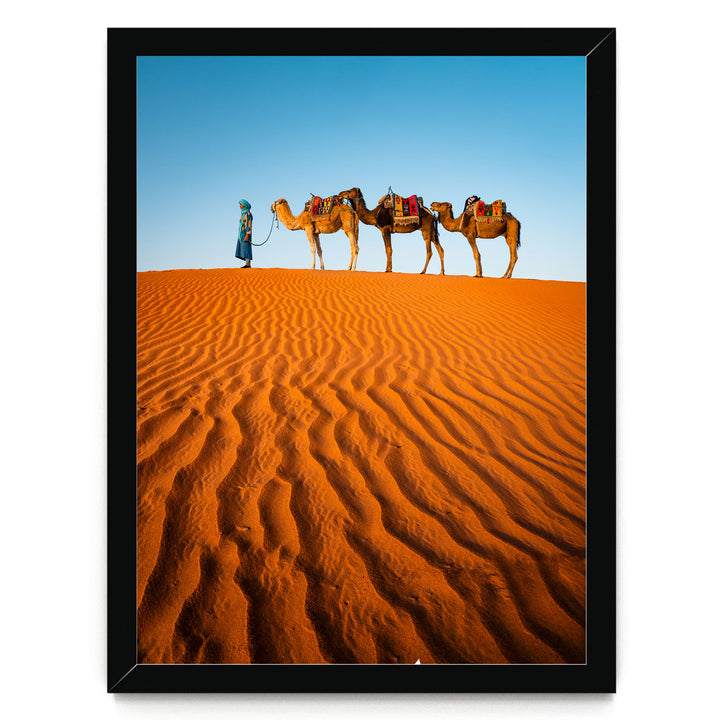 A person in a blue robe leads three camels across a vast, undulating sand dune under a clear blue sky.