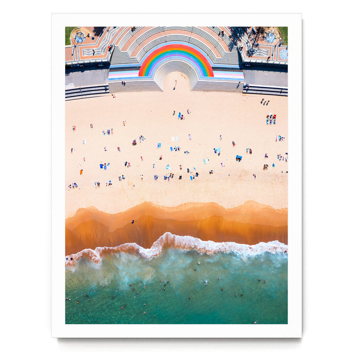 The image is an aerial view of a crowded beach with people enjoying the sun and sand. The beach is lined with a large, colorful arch structure, and the ocean is visible in the background.