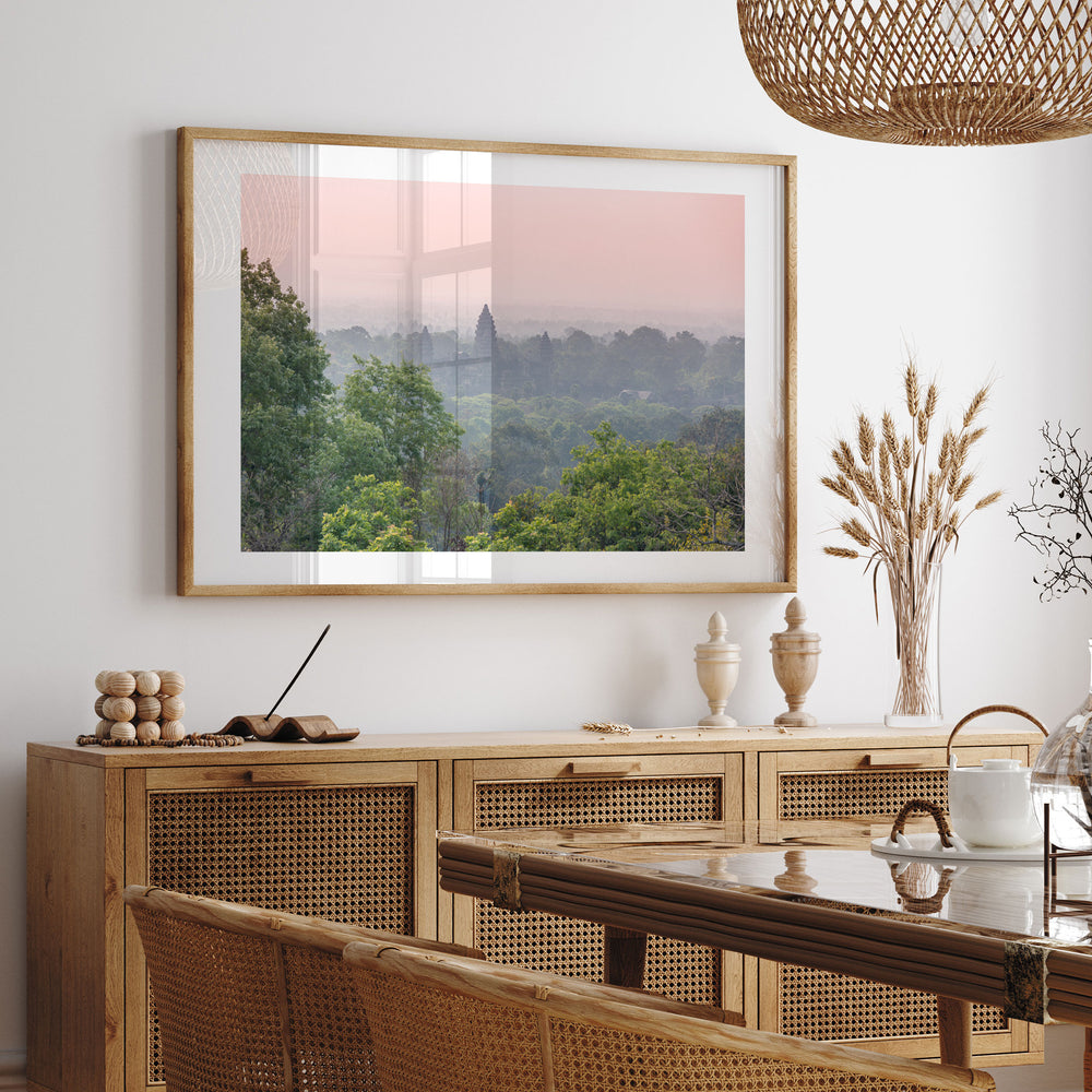 a dining room with a large framed landscape photograph hanging on the wall above a wooden cabinet. The photograph depicts a serene forest scene with a pink and orange sky. The dining table is set with a vase of flowers and a teapot, creating a cozy and inviting atmosphere.