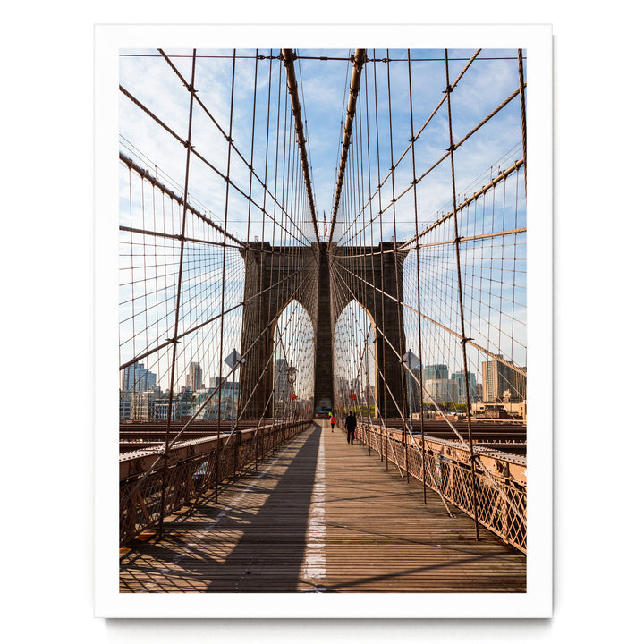 the iconic Brooklyn Bridge, with its distinctive Gothic-style towers and suspension cables, spanning across the frame. The bridge is viewed from the pedestrian walkway, which is lined with wooden planks and metal railings. The scene is set against a backdrop of a clear blue sky and the cityscape of New York City.