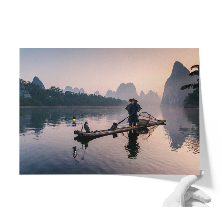 A person stands on a long wooden boat, paddling across a calm body of water surrounded by mountains and trees.