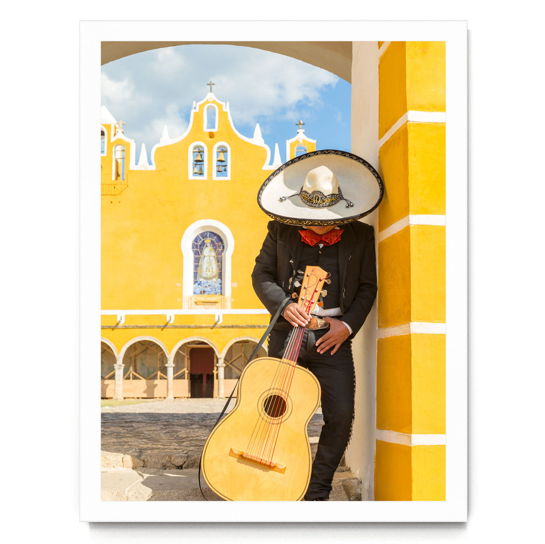 A person dressed in a traditional Mexican outfit, including a sombrero and a black suit, is holding a guitar and leaning against a yellow building with arched windows.