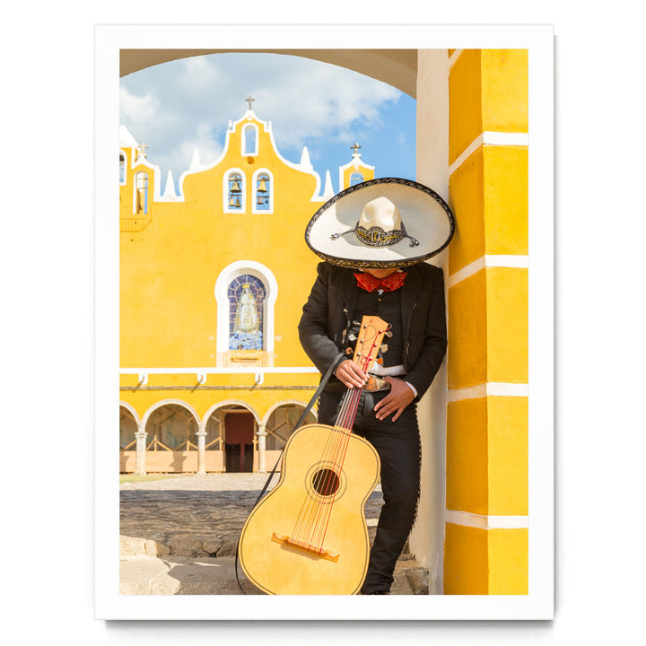 A person dressed in a traditional Mexican outfit, including a sombrero and a black suit, is holding a guitar and leaning against a yellow building with arched windows.