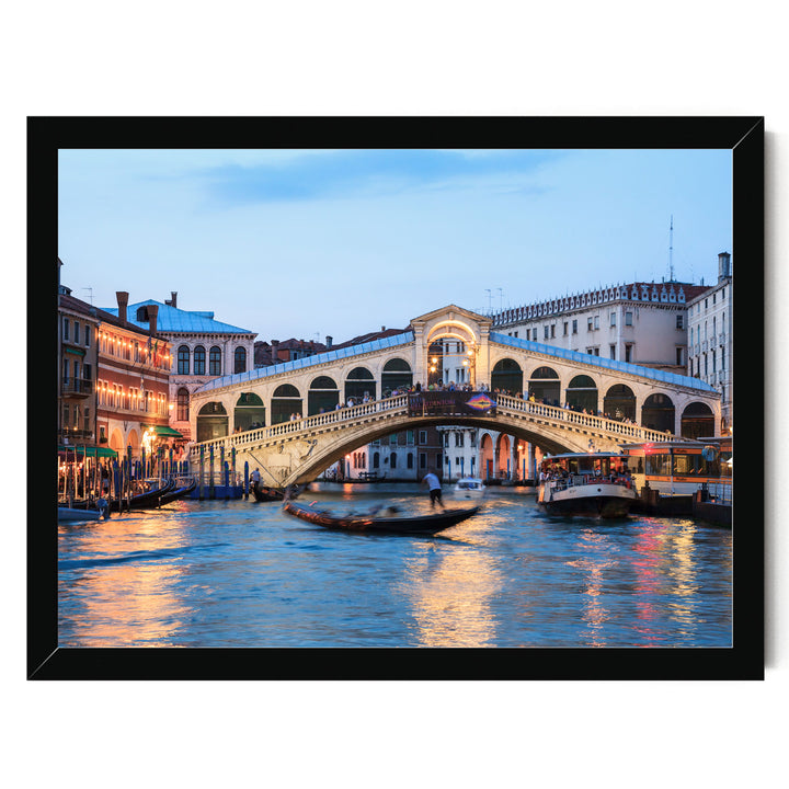 a picturesque scene of the Rialto Bridge in Venice, Italy, with its distinctive white stone arch spanning the Grand Canal. The bridge is illuminated at dusk, creating a warm and inviting atmosphere. The canal is bustling with boats, adding to the lively ambiance of the scene. The image is framed, suggesting it is a piece of art or a photograph.