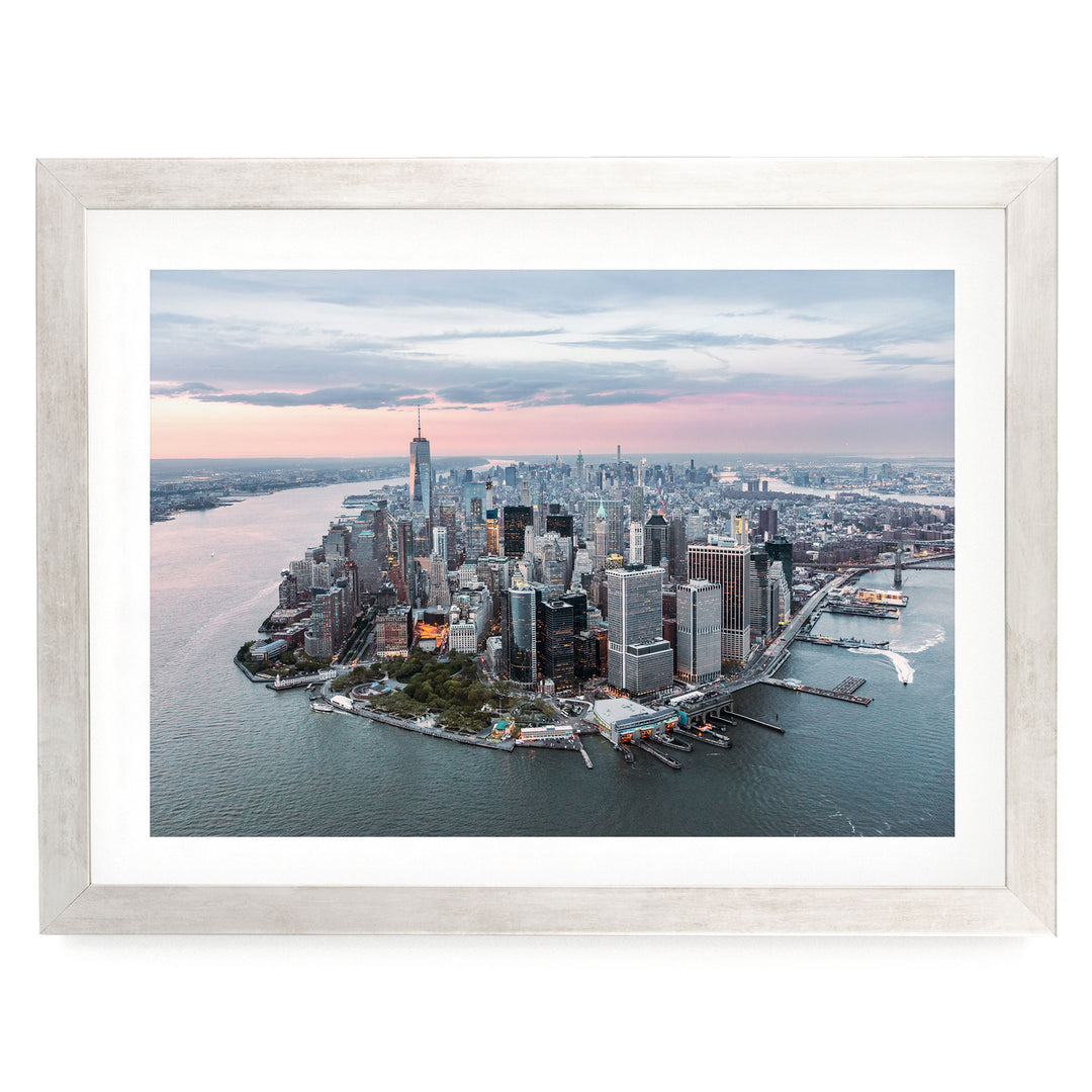The image is an aerial view of a large city, likely New York, with numerous tall buildings and skyscrapers surrounding a body of water. The cityscape is framed within a white border, and the sky above is a mix of pink and blue hues, suggesting either sunrise or sunset.