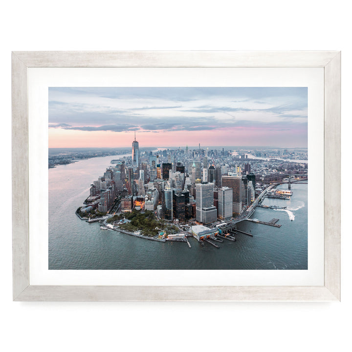 The image is an aerial view of a large city, likely New York, with numerous tall buildings and skyscrapers surrounding a body of water. The cityscape is framed within a white border, and the sky above is a mix of pink and blue hues, suggesting either sunrise or sunset.