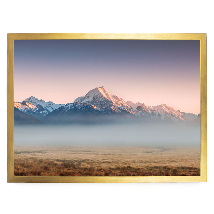 A breathtaking landscape featuring snow-capped mountains in the distance, with a hazy, golden-hued sky above. The mountains are partially obscured by a misty haze, creating a serene and atmospheric scene.
