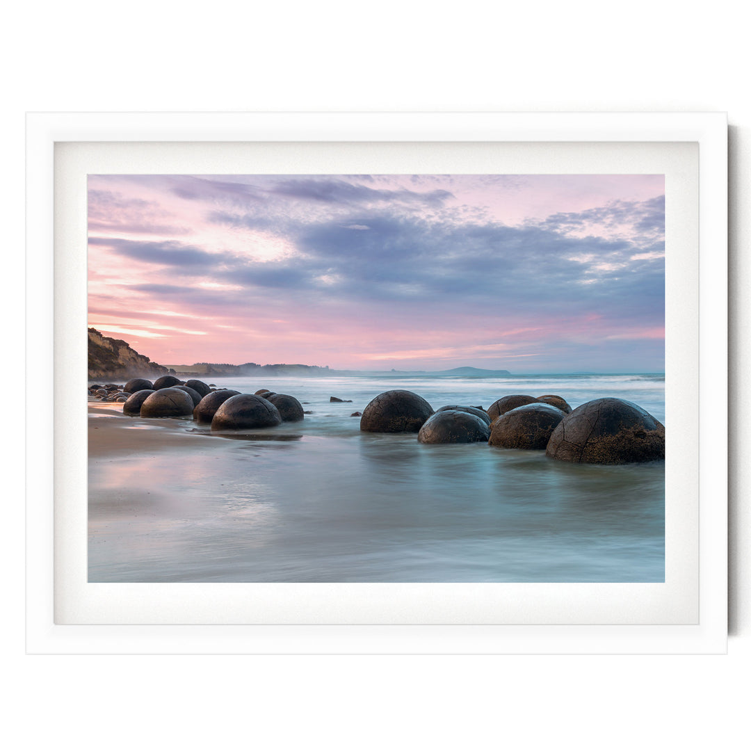 A serene beach scene with a collection of large, round boulders in the water, set against a picturesque sky with pink and purple hues.