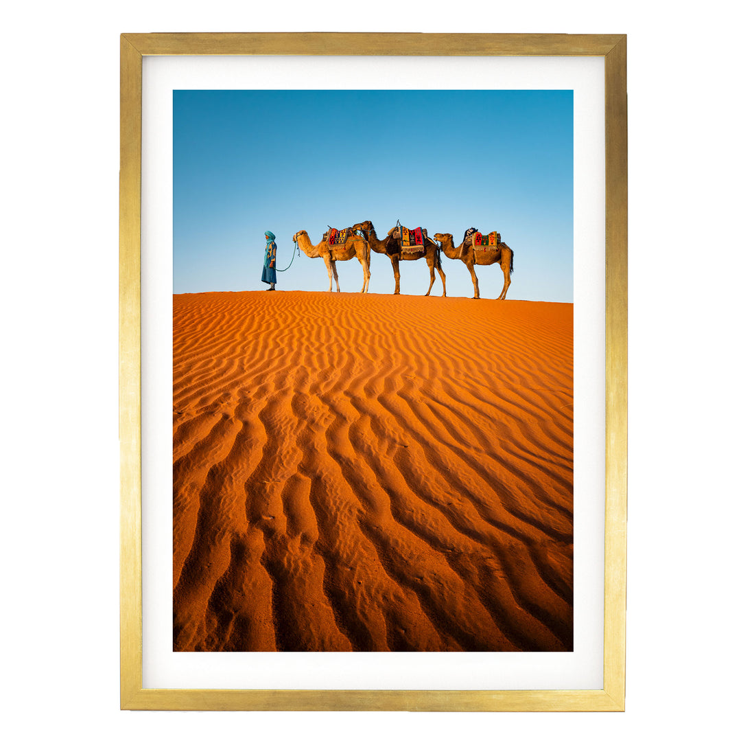 A person in a blue robe leads three camels across a vast, undulating sand dune in a desert landscape.