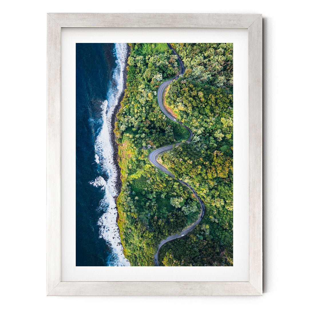 The image is an aerial view of a winding road cutting through a lush, green forest. The road curves gently around the edge of a cliff, with the ocean visible on the left side. The surrounding landscape is dense with trees and vegetation, creating a serene and picturesque scene.
