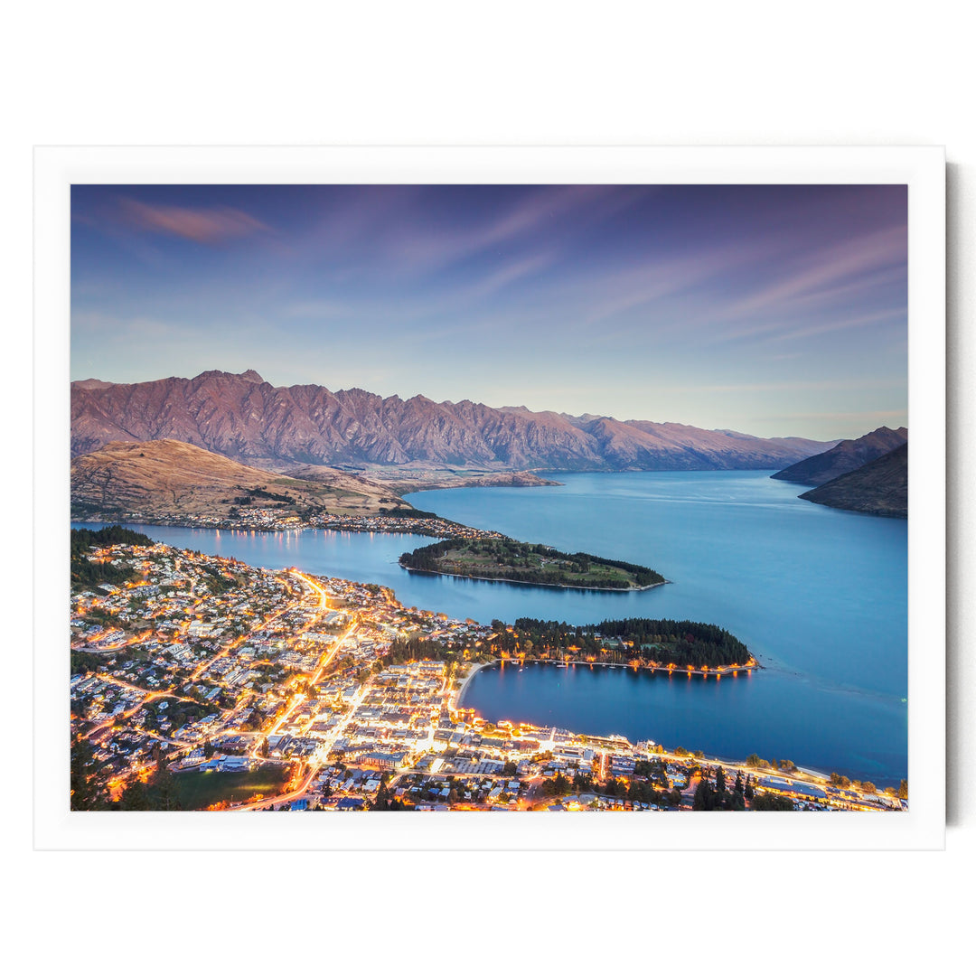 A stunning aerial view of a coastal town nestled between mountains and a deep blue lake, with the town's lights twinkling in the distance against the backdrop of the natural landscape.