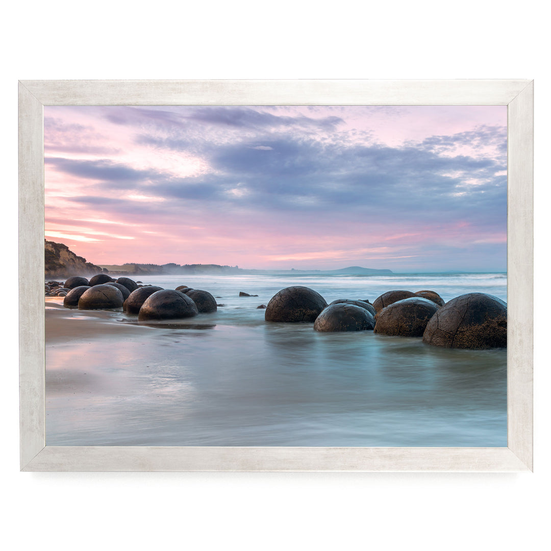 A serene beach scene with a row of large, round boulders in the water, set against a picturesque sunset sky with pink and purple hues.