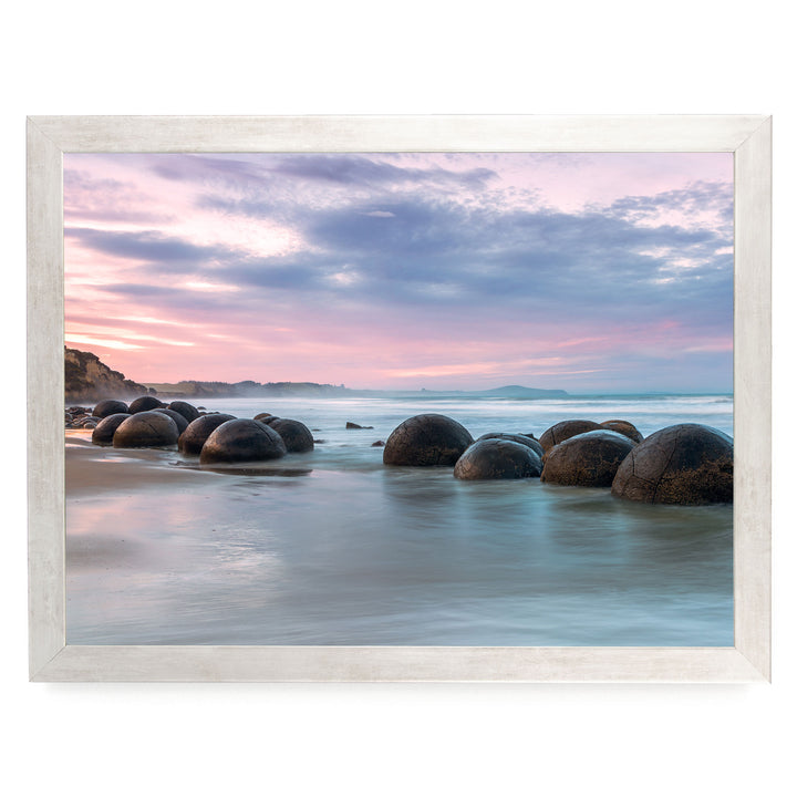 A serene beach scene with a row of large, round boulders in the water, set against a picturesque sunset sky with pink and purple hues.