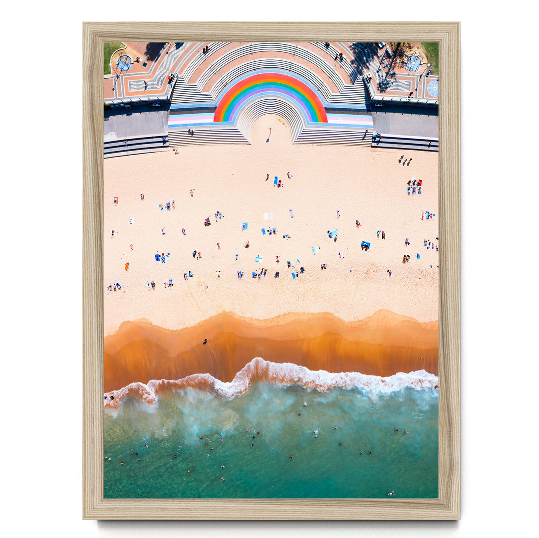 The image is an aerial view of a beach with a large rainbow arching over the top, and a crowd of people enjoying the beach below.