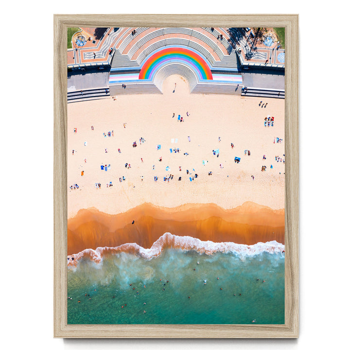 The image is an aerial view of a beach with a large rainbow arching over the top, and a crowd of people enjoying the beach below.